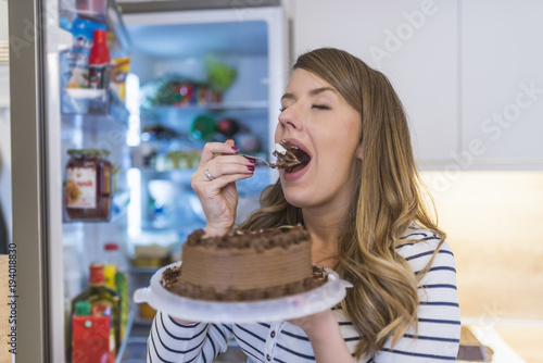 Close-up of young woman standing in front of open refrigerator eating cake
