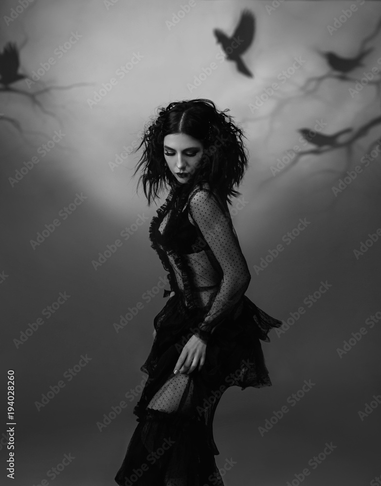 An alien girl in a black, transparent, vintage dress posing against the backdrop of a gloomy forest, which is created by the projector. Artistic black and white photography, play of shadows and light