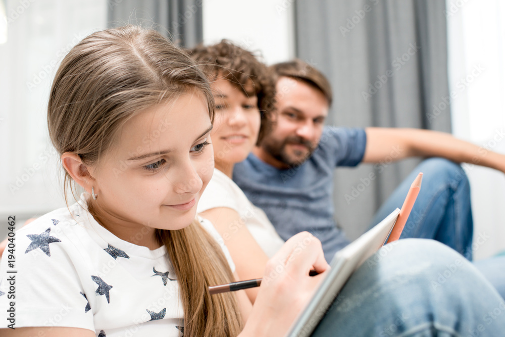 Fototapeta premium Portrait of happy family of three sitting on sofa in living room at home, focus on teenage girl writing notes in foreground