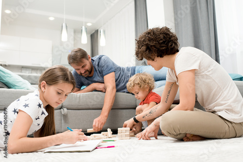 Portrait of happy young family of four enjoying evening together drawing with children on floor in living room