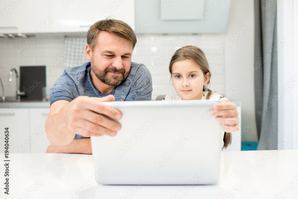 Obraz premium Portrait of smiling bearded man using laptop with daughter sitting at table in modern kitchen