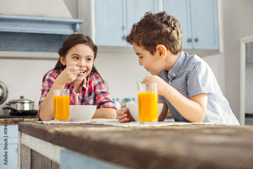 Cheerfulness. Pretty inspired little dark-haired girl smiling and looking at her brother while they having breakfast Cheerfulness. Pretty inspired little dark-haired girl smiling and looking at her brother while they having breakfast
