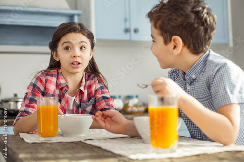 Relationship. Pretty alert little dark-haired girl talking with her brother while they having breakfast and drinking some juice