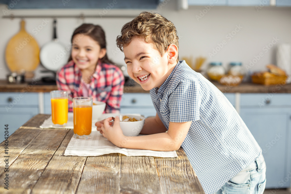 So joyful. Handsome delighted little dark-haired boy laughing and having healthy breakfast with his sister and the girl smiling in the background So joyful. Handsome delighted little dark-haired boy laughing and having healthy breakfast with his sister and the girl smiling in the background