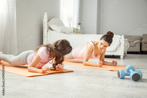 Exercising. Beautiful inspired young dark-haired mother doing some exercises with her daughter while lying on the floor and looking at her girl