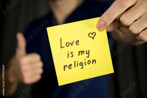 Man holds yellow sticker inscription Love is my religion. Thumb up Blue T-shirt. close-up, selective focus