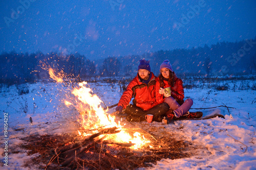 Happy loving couple sitting at a fire in the snow among the fields in the winter            against the background of the forest and the sky on Valentine s Day.
