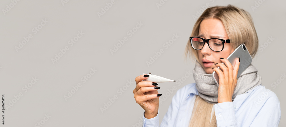 Sick young woman with glasses caught cold, looking at thermometer, talking on mobile phone. Cold, flu season