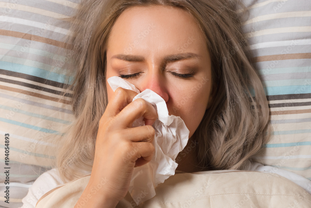 Young European upset and sick female with paper napkin blowing nose, lies on the pillow, closed eyes, indoor/ Sinusitis treatment / Rhinitis, cold, sickness, allergy concept/ Young European upset and sick female with paper napkin blowing nose, lies on the pillow, closed eyes, indoor/ Sinusitis treatment / Rhinitis, cold, sickness, allergy concept/