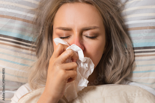 Young European upset and sick female with paper napkin blowing nose, lies on the pillow, closed eyes, indoor/ Sinusitis treatment / Rhinitis, cold, sickness, allergy concept/