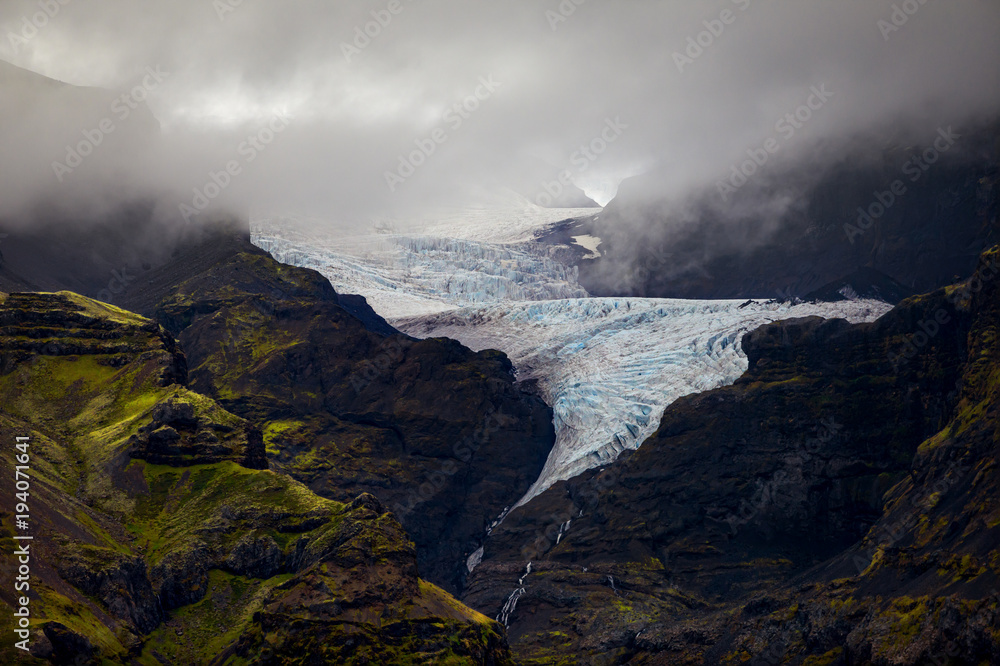 The unique twisting reach of Oraefajok Glacier in Iceland and it reaches down, out of the fog and into waterfalls.  Blue glacier ice is visible, as well as green moss covering rocks of the mountain