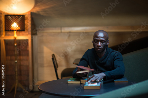 A young Arican man is reading a book in a room at a table. student, businessman