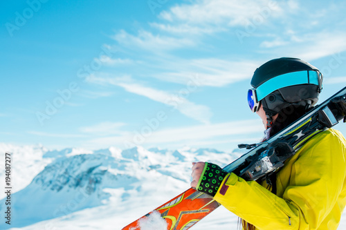 Side view of sporty woman in helmet with skis on her shoulder against background of winter hills