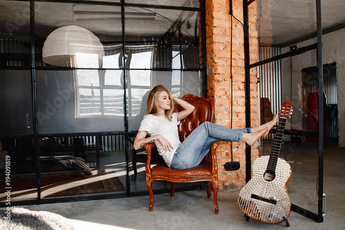 Portrait of the stylish young woman of the blonde in a white t-shirt and jeans in an interior in style the loft. The girl with a guitar.