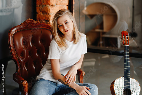 Portrait of the stylish young woman of the blonde in a white t-shirt and jeans in an interior in style the loft. The girl with a guitar.