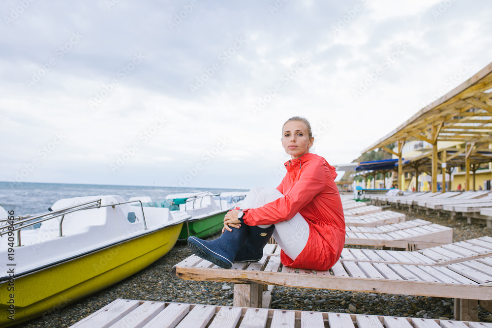 young woman in a red jacket and blue rubber boots sitting on a wooden deck chair near the sea