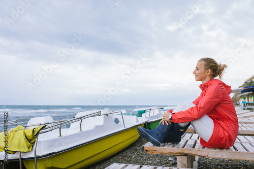 young woman in a red jacket and blue rubber boots sitting on a wooden deck chair and looking at the stormy sea © skvalval