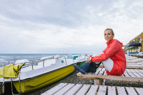 young woman in a red jacket and blue rubber boots sitting on a wooden deck chair near the sea
