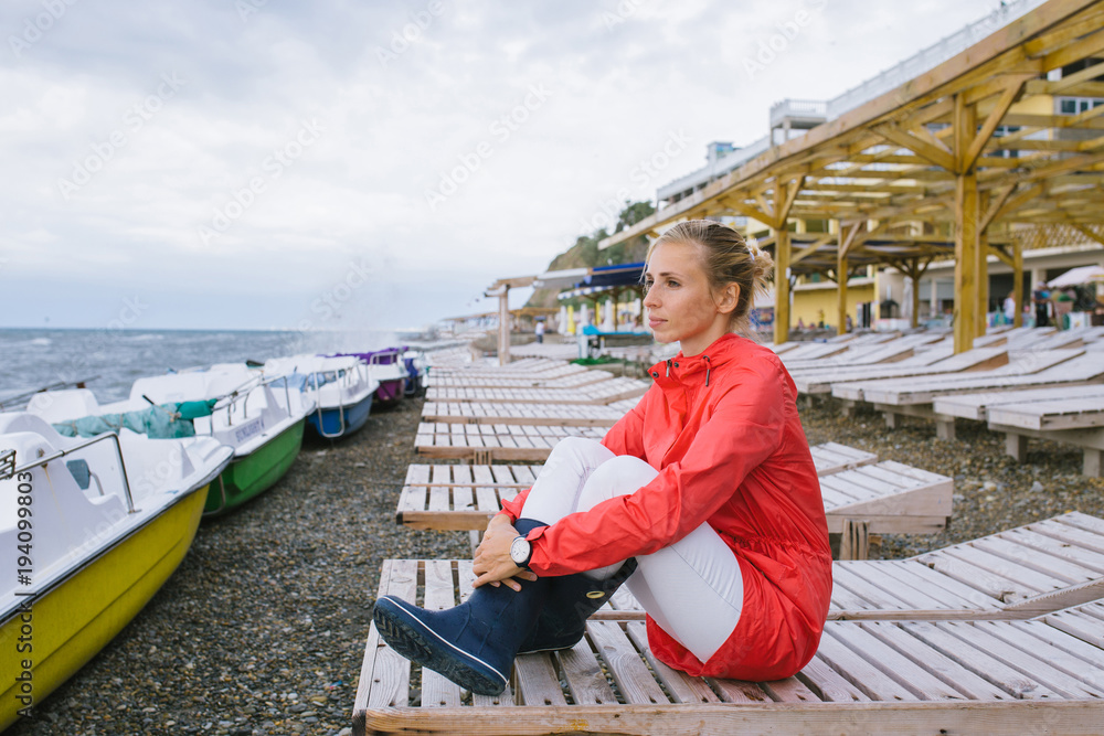 young woman in a red raincoat and blue rubber boots sitting on a wooden deck chair and looking at the stormy sea
