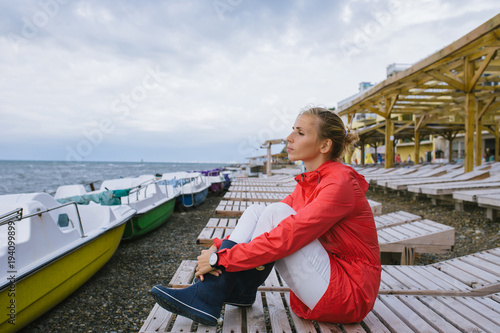 young woman in a red raincoat and blue rubber boots sitting on a wooden deck chair and looking at the stormy sea