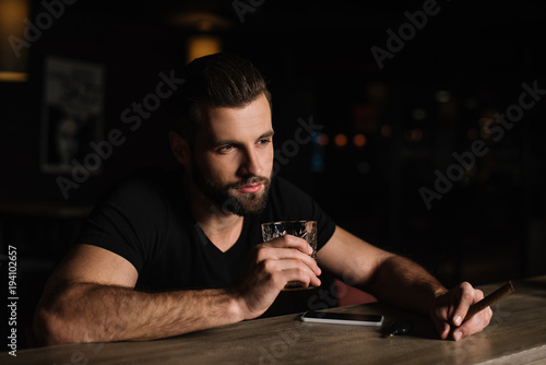 handsome visitor sitting at bar counter and holding glass of whiskey and cigar