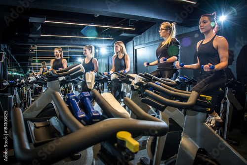 Group of young sporty women working out with dumbbells on cycling class at gym.