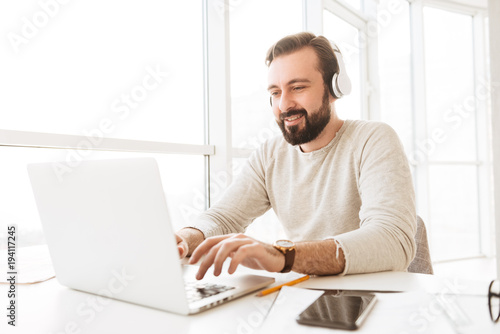 Positive european man with short brown hair chatting or scrolling social network on laptop, while listening to music via headphones