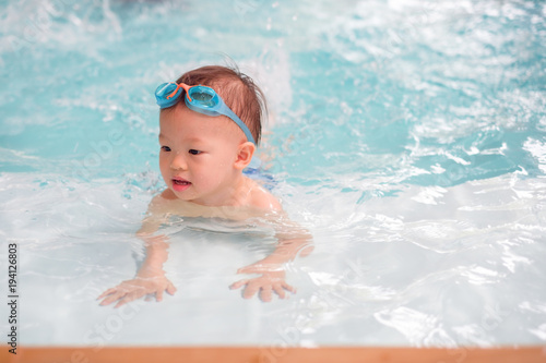 Cute little Asian 18 months / 1 year old toddler boy child in trunks wear swimming goggles learn to swim at indoor pool, Swimming school for small children. Happy kid enjoy active lifestyle concept