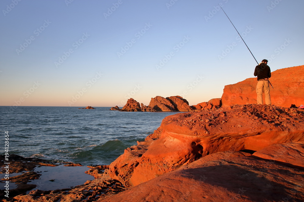 Nadmorski krajobraz, skalisty brzeg, na pięknej brązowoczerwonej skale stoi wędkarz, tyłem, z wędką, w tle morze i inne skały w pięknym świetle zachodzącego słońca - obrazy, fototapety, plakaty 