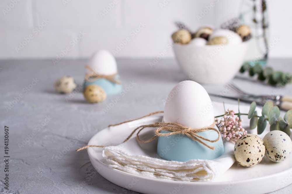 Fototapeta premium Easter festive table setting with white chicken eggs in eggs cups, leaf sprigs of eucalyptus. On a gray concrete background.