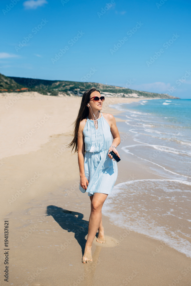 Girl with long hair in sunglass and sky blue dress is slowly walking on the beach and listening music on earphones