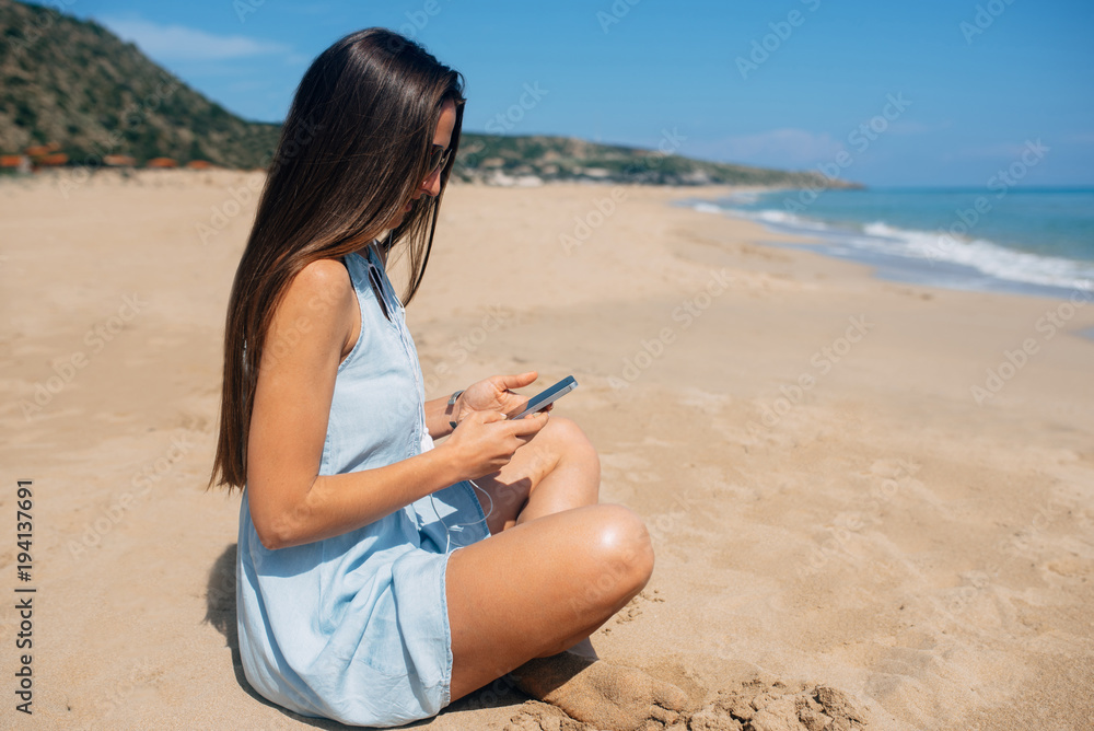 Brunette in sky blue dress with long brown hair sits in lotus pose on the sand and looks something in her cell phone