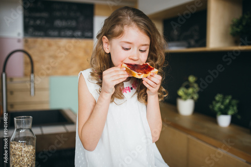 Girl sitting on the table in the kitchen and eating bread and jam 
