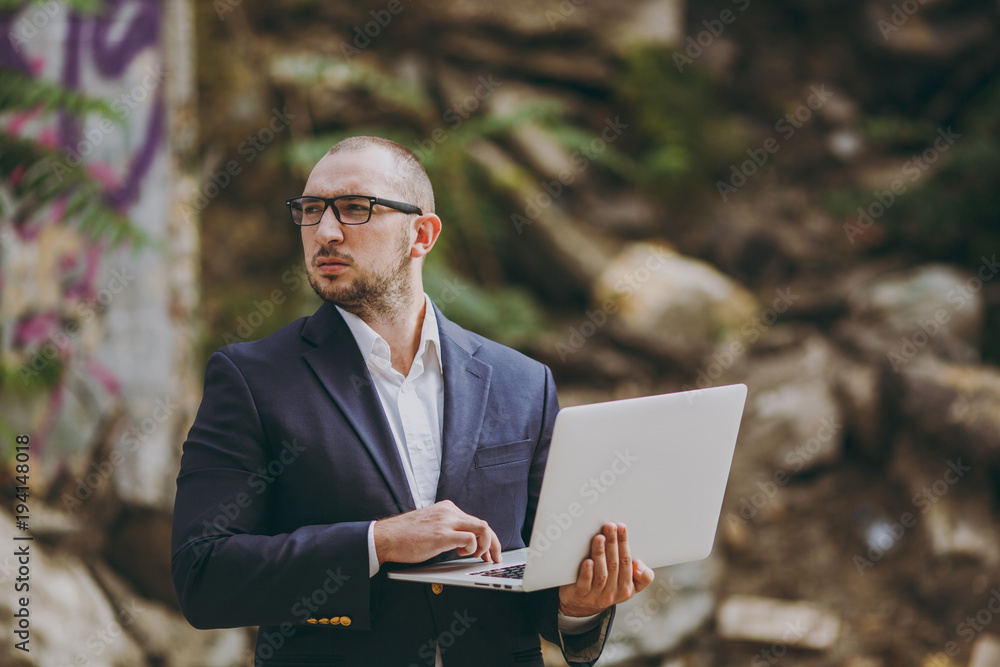 Young successful smart businessman in white shirt, classic suit, glasses. Man standing and working on laptop pc computer near ruins, debris, stone building outdoors. Mobile Office, business concept. Young successful smart businessman in white shirt, classic suit, glasses. Man standing and working on laptop pc computer near ruins, debris, stone building outdoors. Mobile Office, business concept.