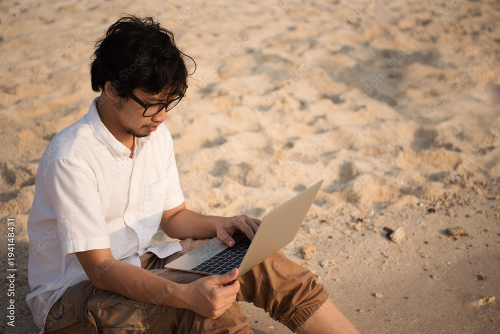 Asian man with his computer on the beach with waves from sea and golden sunlight at evening. working on computer at outdoors while traveling at sea in summer holiday. freelancer work concept.