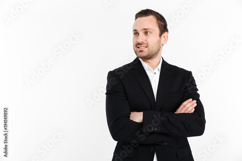 Portrait of young businessman looking aside while standing in suit with broad smile keeping arms folded, isolated over white background