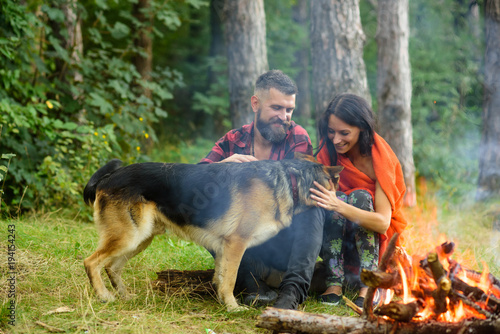 Romantic couple with dog sitting near bonfire, green forest background.