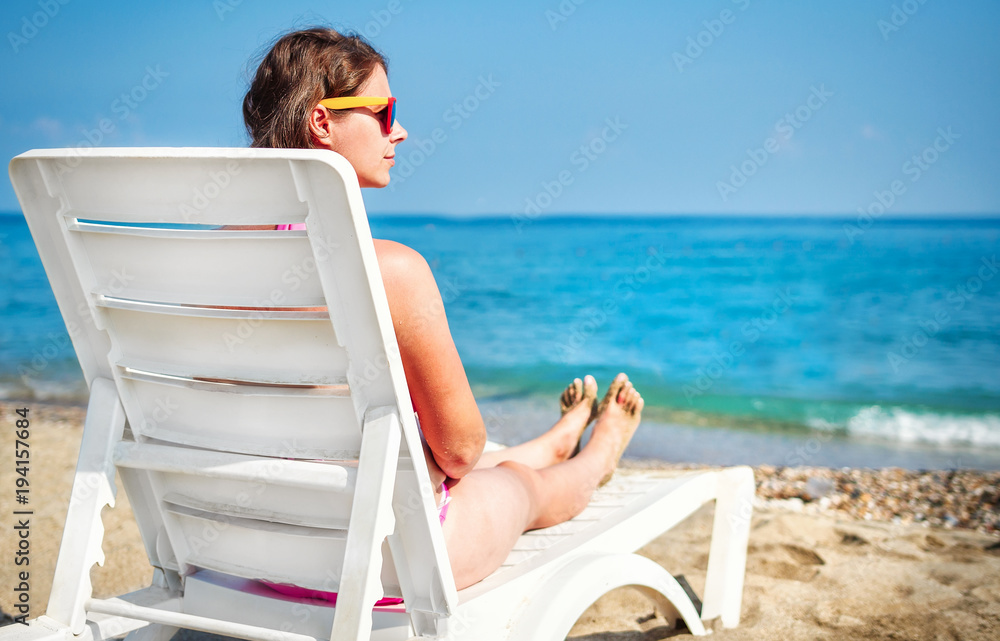 Beautiful young girl on sea beach sitting on sun lounger sunbathing. Attractive woman in glasses sunbathing near the sea. Relax in a tropical resort. Summer vacation on the beach
