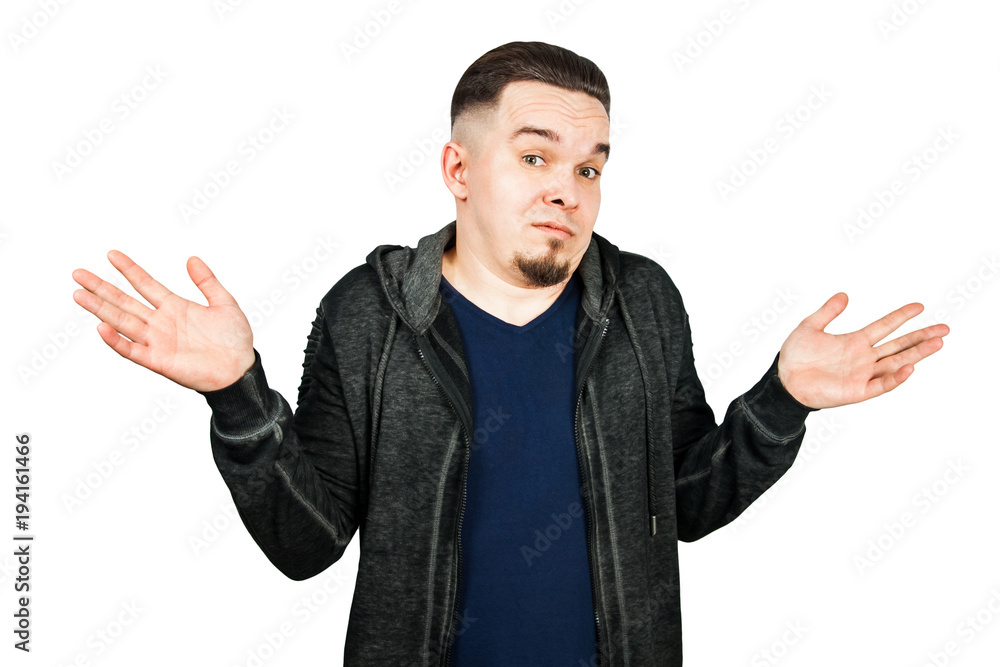 Fototapeta premium Doubtful bearded guy shrugs shoulders in bewilderment, tries to make a decision, isolated over white concrete wall. Hesitation and uncertainty
