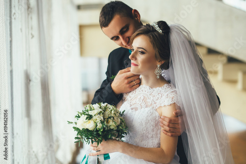 Fotografija Young groom hugs bride tender standing in the hall before a window