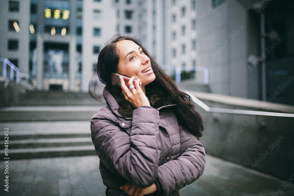 young girl talking on mobile phone in courtyard business center. girl with long dark hair dressed in winter jacket in cold weather speaks on phone on background buildings made of glass and concrete young girl talking on mobile phone in courtyard business center. girl with long dark hair dressed in winter jacket in cold weather speaks on phone on background buildings made of glass and concrete