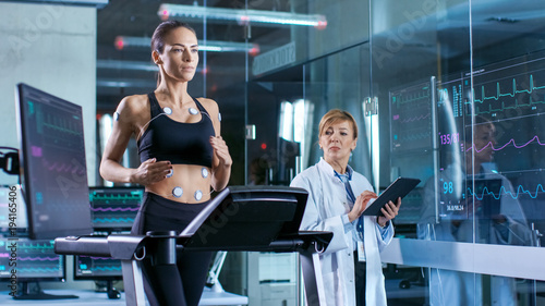 Foto Beautiful Woman Athlete Runs on a Treadmill with Electrodes Attached to Her Body, Female Physician Uses Tablet Computer and Controls EKG Data Showing on Laboratory Monitors