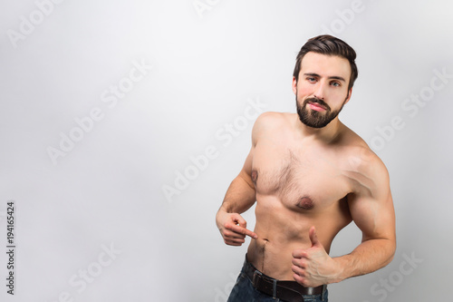Good-looking and awesome guy is standing of a wall and pointing on himself and how his body is good. Young man is self-confident and loves himself quite much. Isolated on white background.