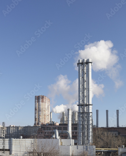 White smoke comes from a white chimney heat station pipe on a blue sky background.