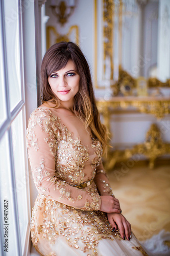 Portrait of cute girl in gold dress, sits on window sill, in luxurious room