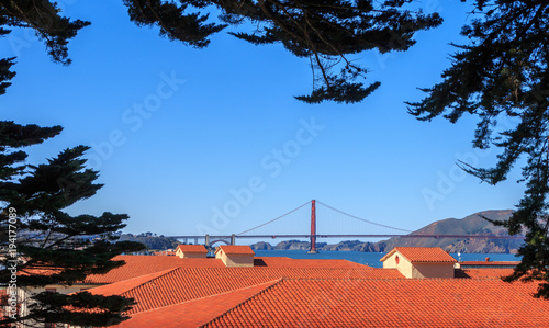 A view of the Golden Gate Bridge from the heights above Fort Mason in San Francisco. Trees frame the picture of red tile roofs and a view of the bay. A blue sky is in the background. photo