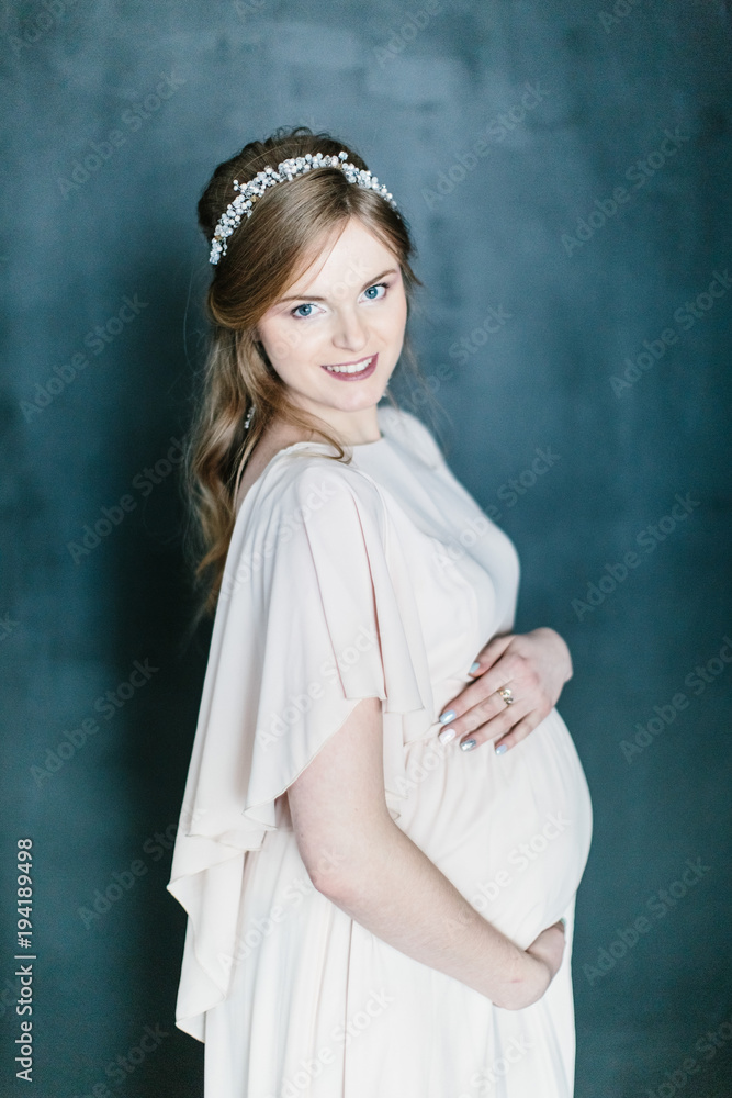 Young beautiful pregnant woman with a beautiful diadem in the hair poses standing on dark background. Young beautiful pregnant woman with a beautiful diadem in the hair poses standing on dark background.