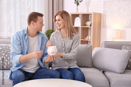 Young couple putting coin into piggy bank at home