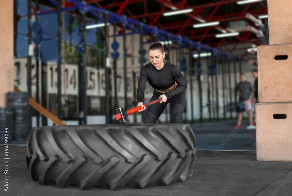 Fototapeta premium Young muscular woman hitting heavy tire with hammer in gym