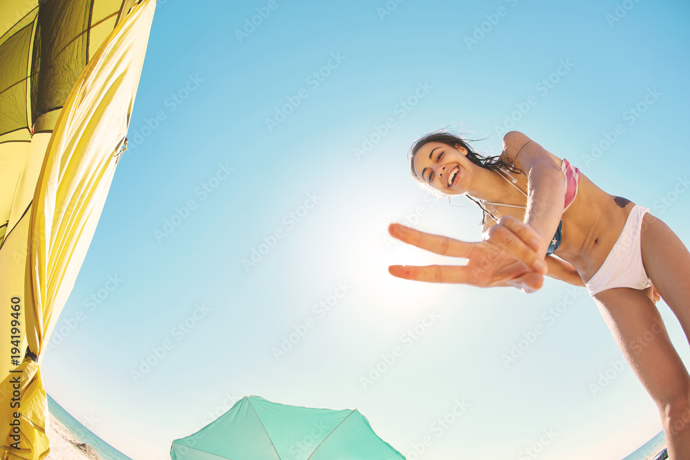 Fototapeta premium beautiful smiling woman looks inside the tent and shows Victory sign in camera . view from inside tent on the sea and sandy beach with beach umbrella and beach chair on a bright sunny day. young